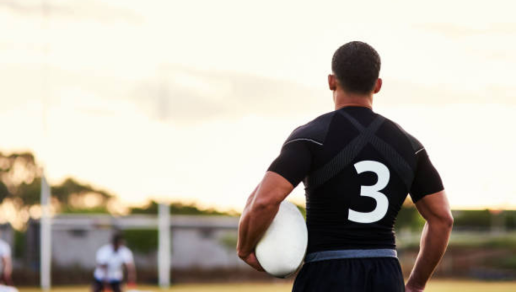 A rugby player wearing a black jersey with a large number 3 on his back, holding a rugby ball while looking out at a field during sunset.