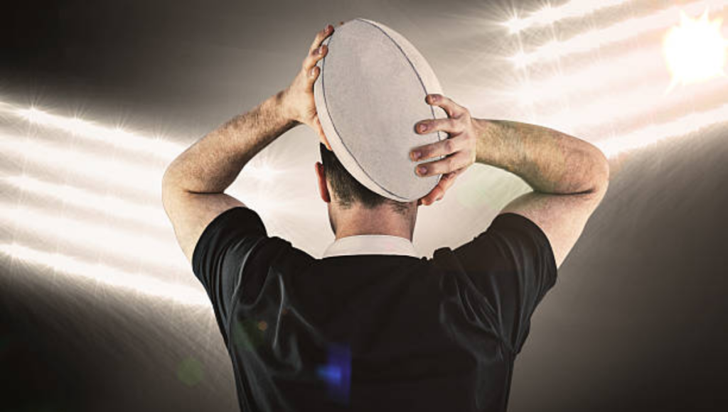 The back view of a rugby player holding a rugby ball above their head, facing bright stadium lights, ready to throw the ball.