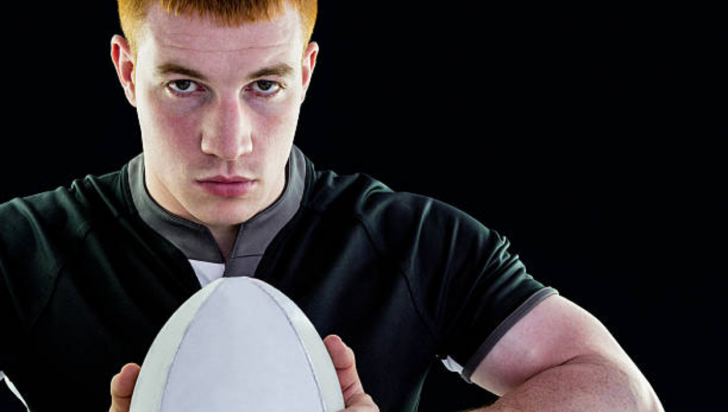 A close-up of a rugby player with short red hair, wearing a black shirt, intensely holding a rugby ball with both hands in front of his chest against a black background.