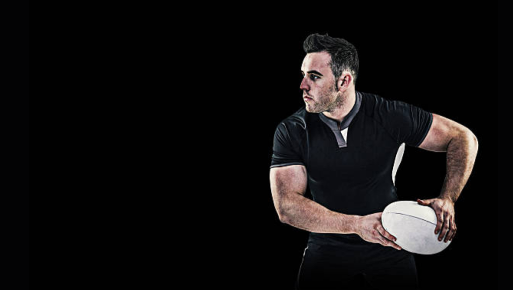 A muscular rugby player in a black shirt holding a rugby ball while preparing to pass it, set against a dark background.