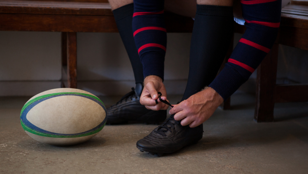 A rugby player lacing up their boots, getting ready for the game with a ball next to them.