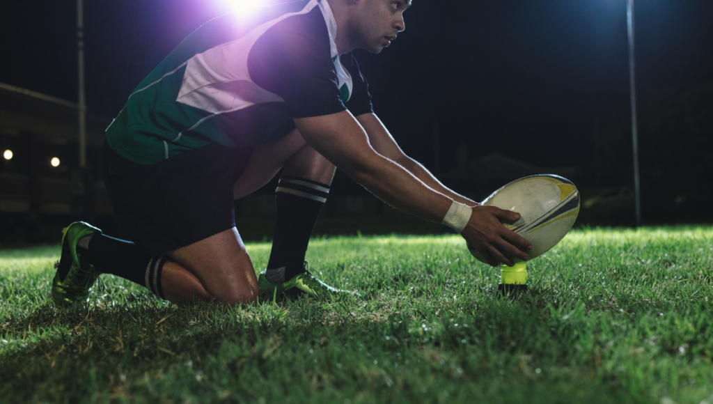 A rugby player in action at night, placing the rugby ball on a kicking tee on a grass field under bright stadium lights.