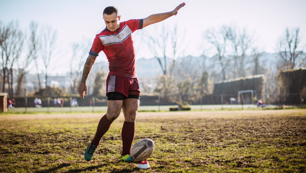 A rugby player in a red uniform kicking a ball placed on a tee on a grassy field during practice.