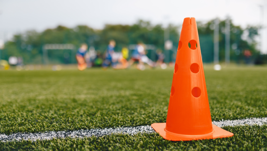 A close-up of an orange rugby training cone placed on a grass field, with blurred players and equipment in the background.