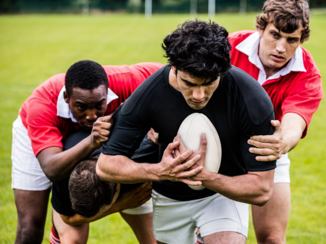 A group of rugby players engaged in a close tackle on the field, with one player holding the rugby ball tightly as others try to stop him.