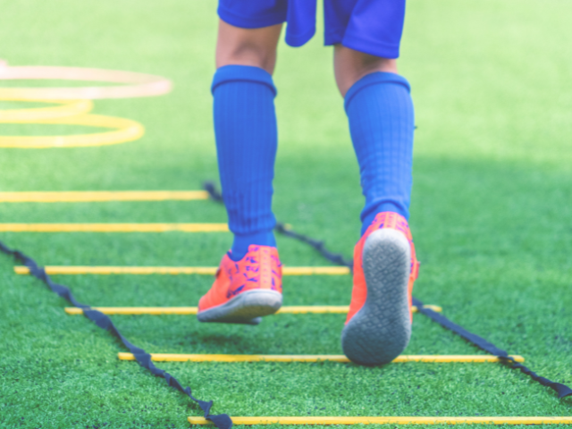 A young athlete wearing orange rugby boots, training on artificial turf with a ladder drill.