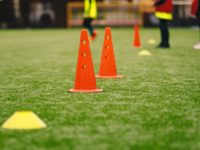 Orange rugby cones with holes placed on a grassy field during a training session with players visible in the background.