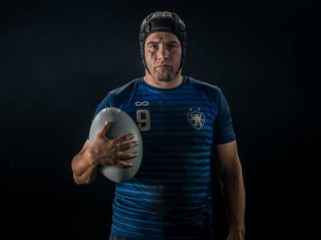 A rugby player wearing a dark blue and black shirt holding a rugby ball, looking serious, standing in a dark background.