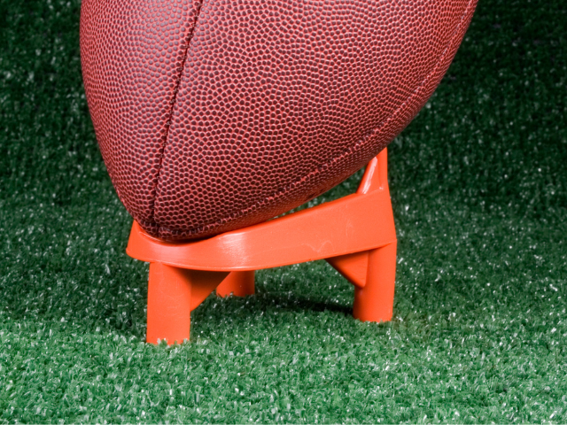 A football resting on a red plastic kicking tee, positioned on a turf field.