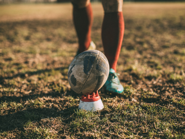 A worn rugby ball set on a kicking tee, with a player's legs visible in the background on a dirt field.