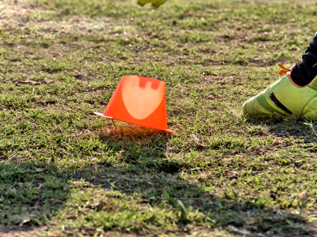 A close-up of an orange marker cone knocked over on a grassy field with a player's foot visible in the background.