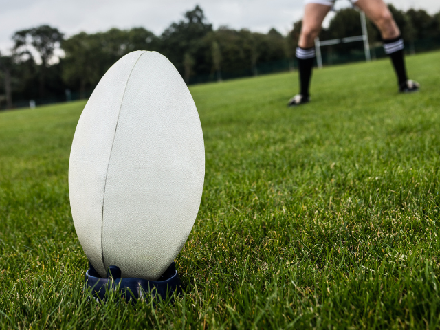 A rugby ball placed on a dark blue kicking tee on a green field with a player in the distance preparing for a kick.