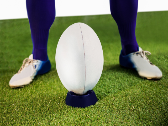 A close-up of a rugby ball placed on a dark blue kicking tee on green grass with a player’s feet visible in the background.