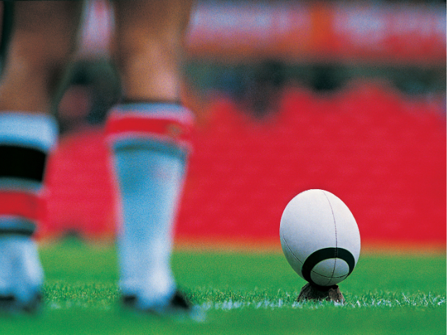 A rugby kicking tee holds a ball upright on a synthetic turf, ready for a player to strike.