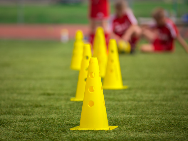 Yellow training cones lined up on a grassy field with players wearing red uniforms in the background.