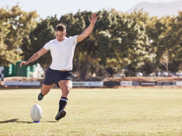 Rugby player kicking a ball off a tee in a sunny outdoor field.