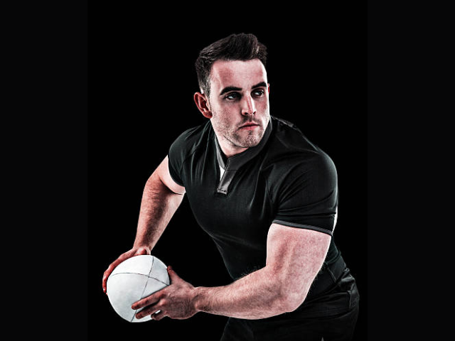 A rugby player in a black jersey holding a rugby ball, preparing to pass it, with an intense expression, set against a dark background.