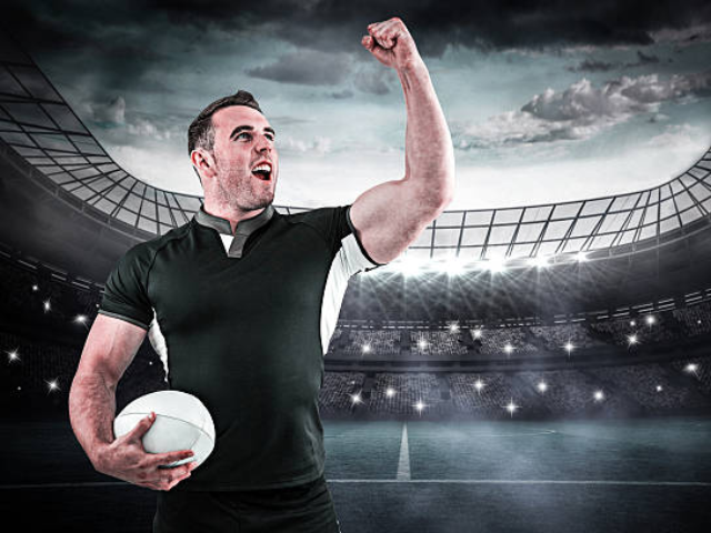 A rugby player in a green and black jersey holding a rugby ball, raising his fist in celebration on a stadium field with bright lights and a cloudy sky.