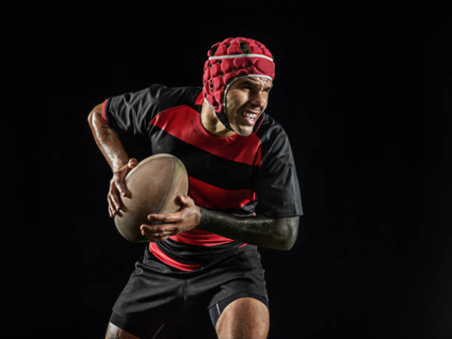 A rugby player in a red and black jersey with a red protective helmet, holding a rugby ball in an action pose on a black background.