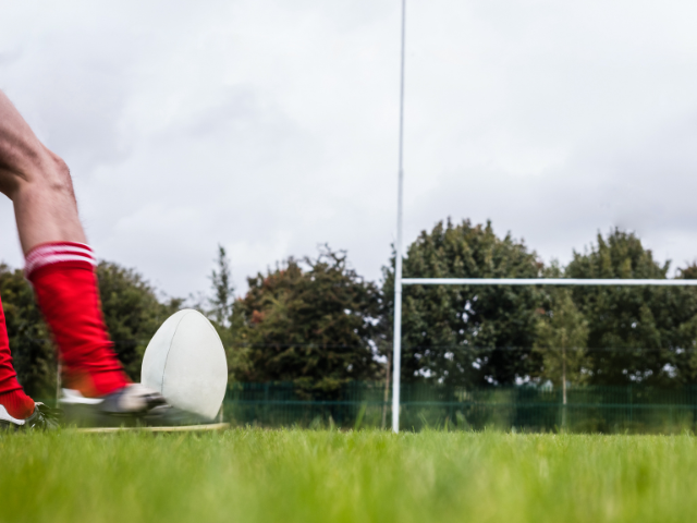 A rugby ball positioned on a tee, ready for a kick as a player in red socks approaches.