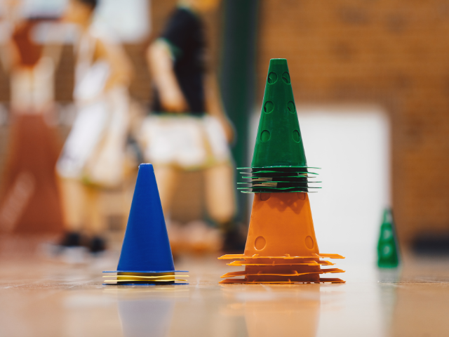 Stacked green, orange, and blue training cones of different sizes set up on an indoor court with players in the background.