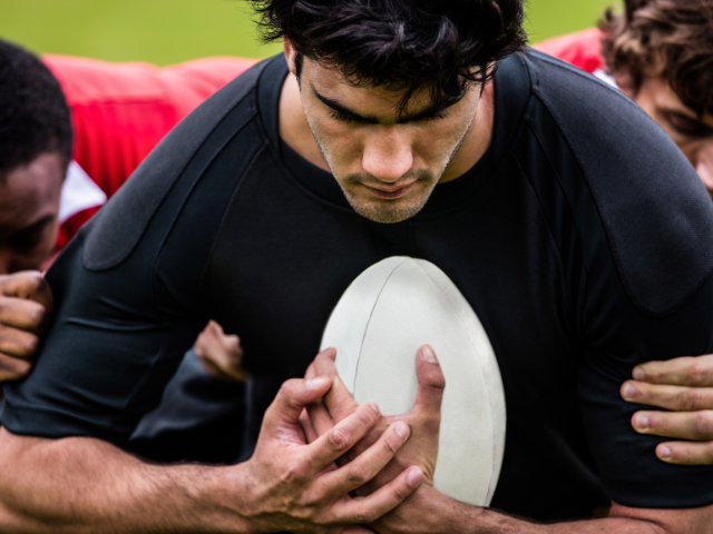 Rugby players in a close-up scrum, focusing on one player's intense grip on the rugby ball as his teammates provide strong support from behind.