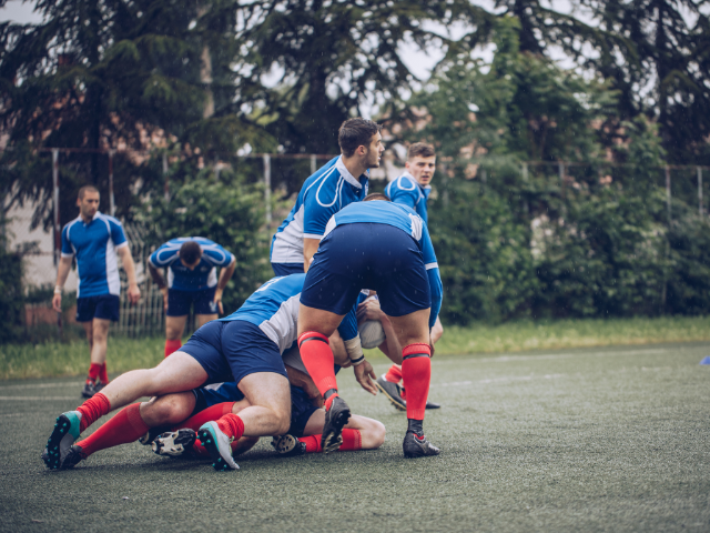 A group of rugby players in blue and red uniforms competing in a tackle, with two players bent over, securing the ball while teammates are ready to support and push forward.