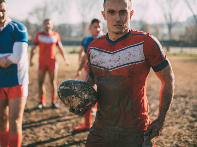 A rugby player holding a muddy rugby ball after a tough game, with other players standing in the background.