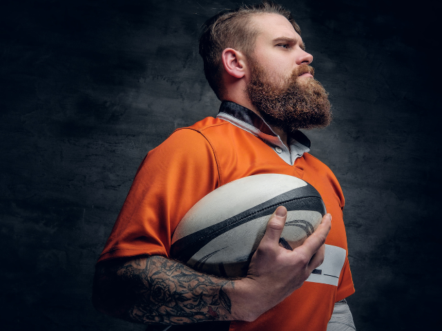 A bearded rugby player confidently holds a rugby ball, ready for action, showcasing the grit and determination required in the sport.