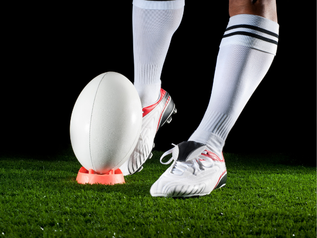 A close-up view of a rugby ball placed on an orange kicking tee, set against a background of lush green grass and a night sky.