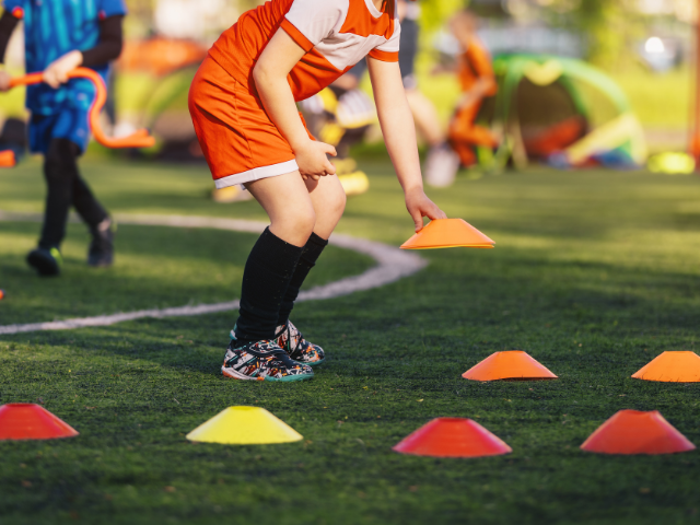 A young player in an orange uniform picking up a training marker during a practice session on a grass field.