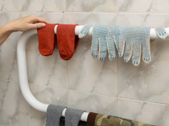 Two red socks and a pair of gloves with blue dotted grips are hanging over a drying rack, set against a tiled bathroom wall.