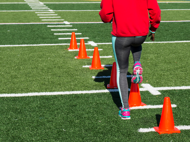 A person in running shoes and leggings stepping through a row of orange cones during an outdoor training session on a field with yard lines.