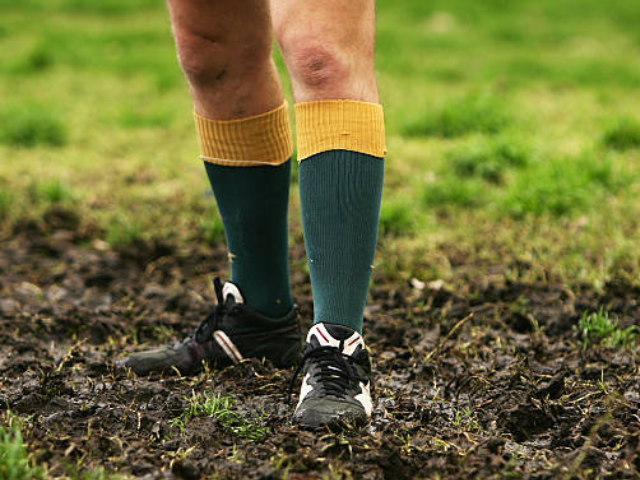 A person standing on muddy ground, wearing green socks with yellow tops and black sports shoes.