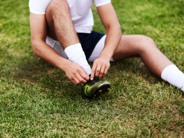 A person sitting on the grass, wearing white socks, tying their black and green sports shoes.