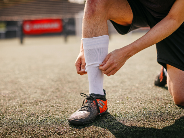 A soccer player on an artificial grass field adjusting his white knee-high socks while wearing black and orange cleats.