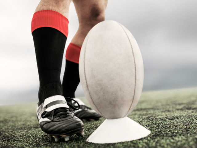 A close-up of a rugby player preparing to kick a ball, wearing black cleats and red socks, standing on a grassy field with a white rugby ball on a tee.