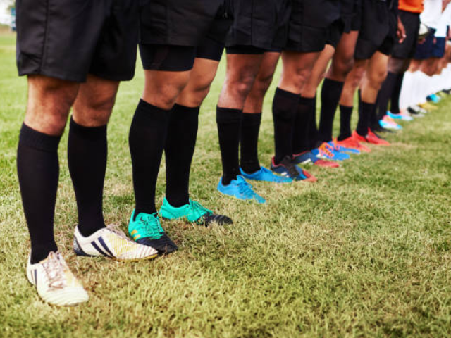 A row of rugby players standing side by side, each wearing different colored cleats and black socks on a grassy field.