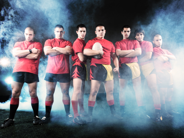 A group of rugby players standing in a foggy, intense atmosphere, all dressed in red shirts and black or yellow shorts, looking serious and determined.