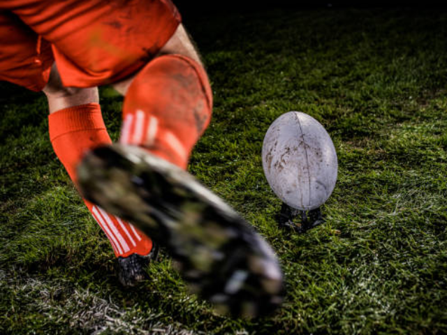 A rugby player in an orange uniform kicking a muddy rugby ball from a stand during a game.