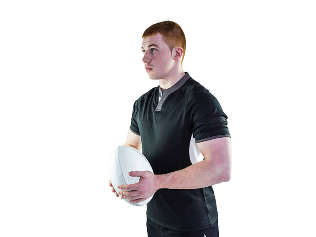 A rugby player with red hair standing in a black sports shirt, holding a rugby ball with both hands against a plain white background.