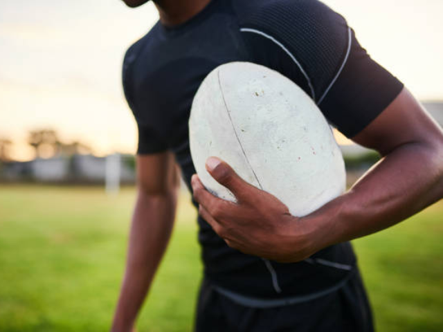 A rugby player in a black sports shirt holding a white rugby ball under his arm on a grassy field, with the sun setting in the background.