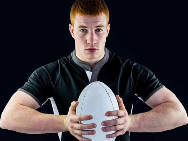 A rugby player in a black uniform holding a rugby ball, standing in a ready position, against a black background.