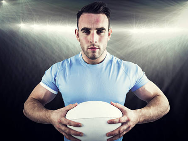 A determined rugby player in a light blue jersey, holding a rugby ball and standing under bright stadium lights, ready for action.