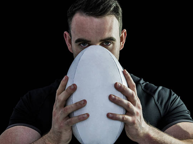 A rugby player in a black uniform holding a white rugby ball, standing against a dark background with a focused expression.