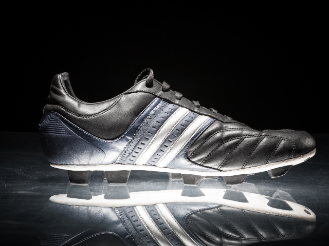 A black rugby boot with white and silver accents, reflecting on a glossy surface.