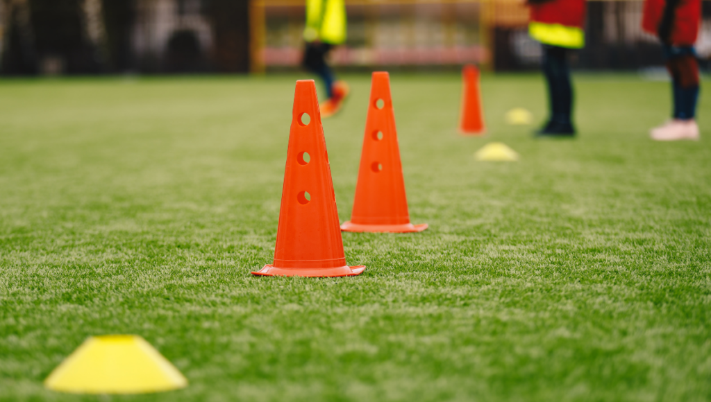 Orange rugby cones with holes lined up on a grassy field with blurred figures and cones in the background during a training session.