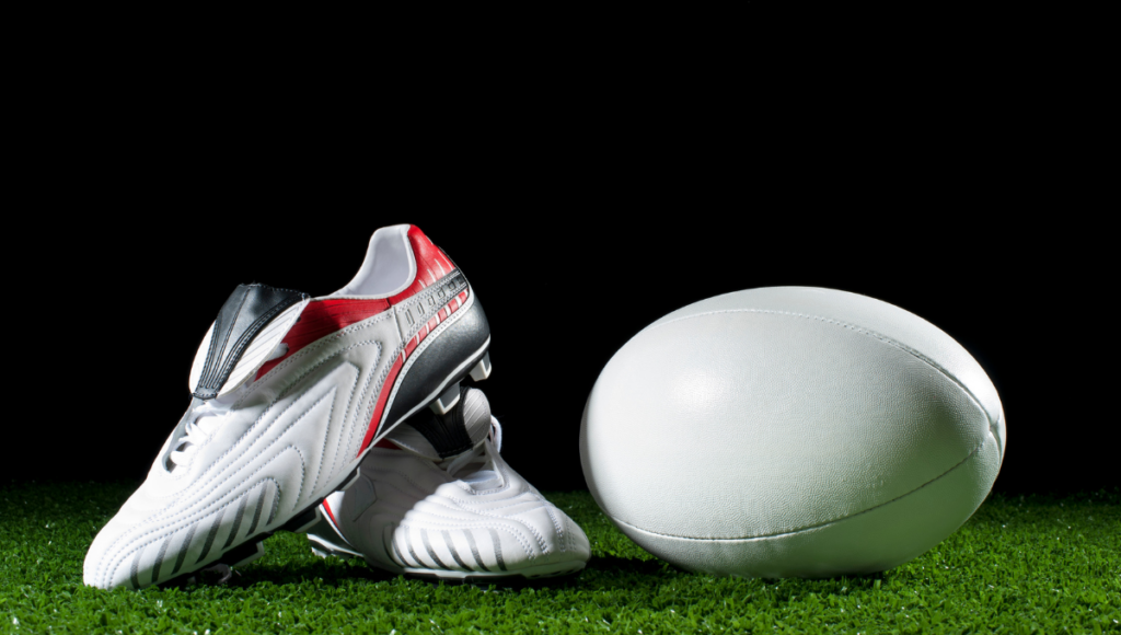 Red and white rugby boots paired with a white rugby ball on green turf.