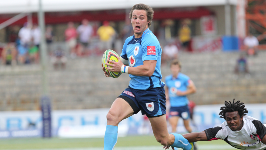 A rugby player in blue uniform running with the ball, while a defender dives to tackle him.