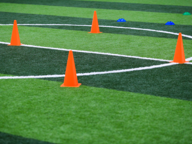 Four orange cones placed in a square formation on an artificial turf field with white lines marking the surface.